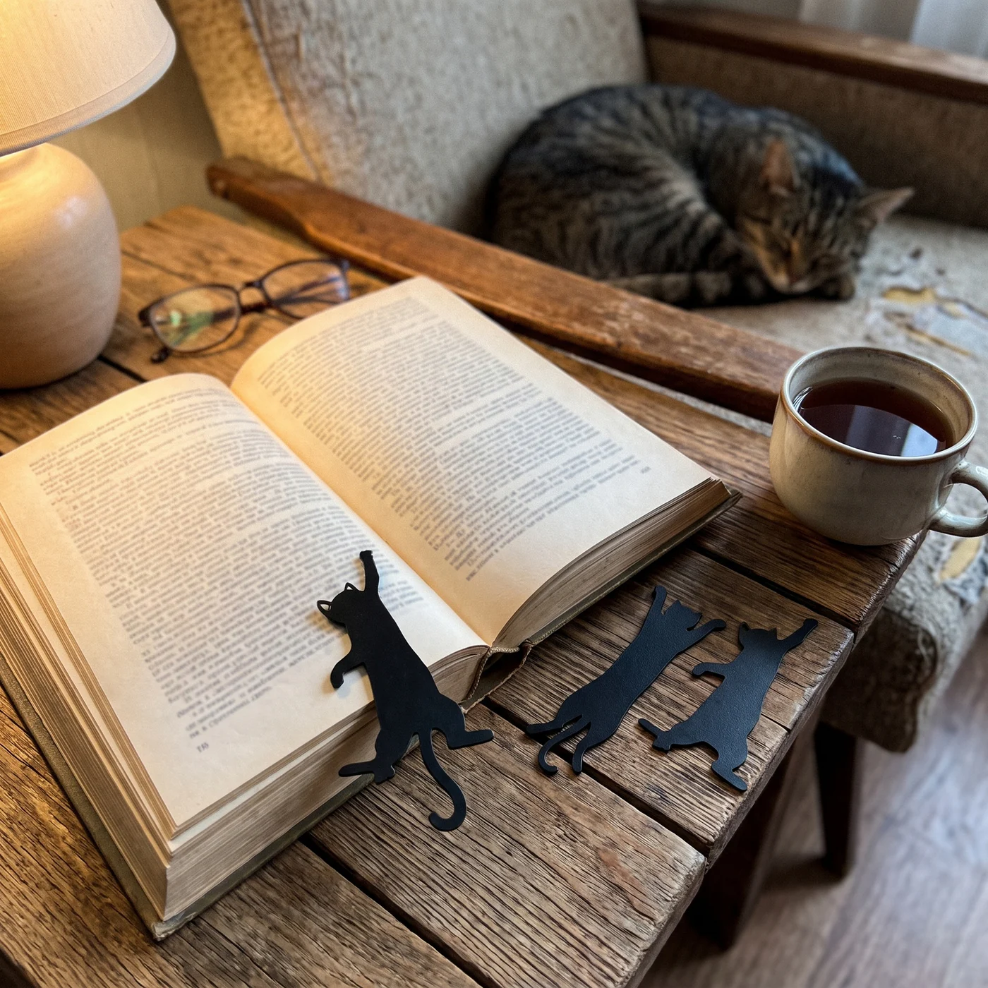 Black metal cat bookmarks from The Bookmark Cats on an open book beside a cup of tea, with a tabby cat asleep behind
