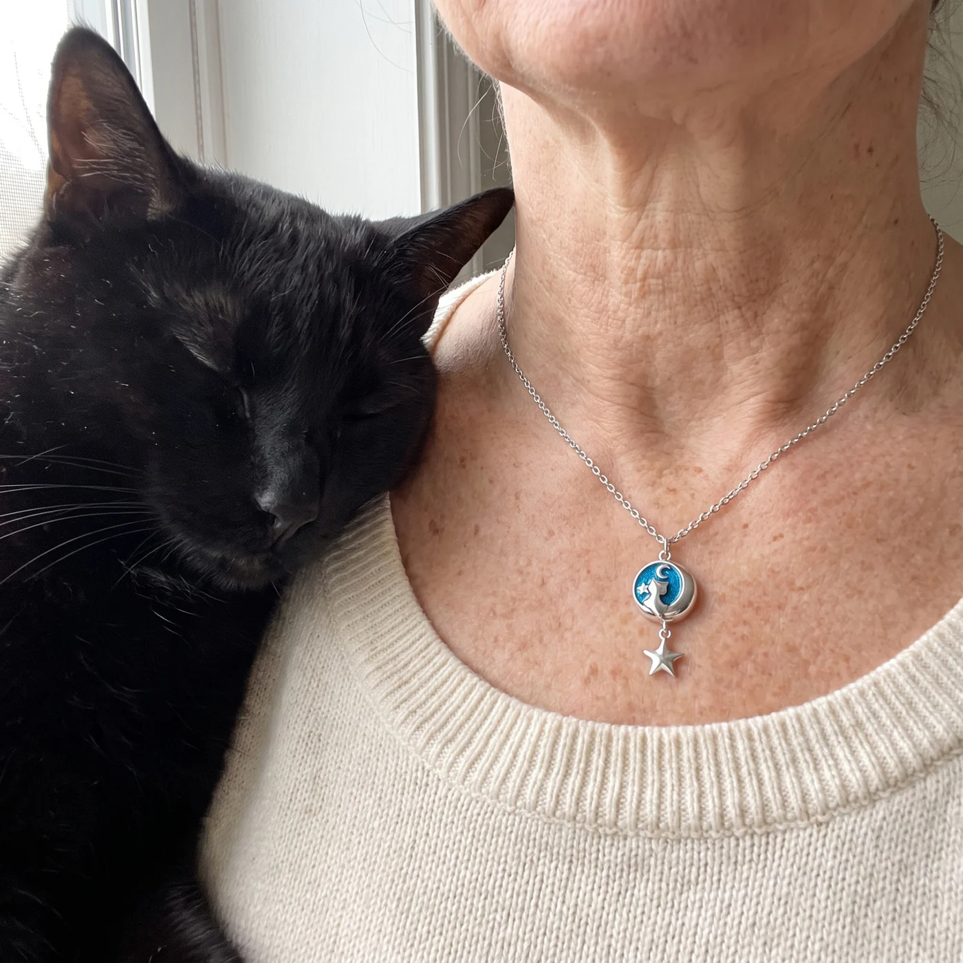 The Moon Cat Necklace worn with a cream sweater: a silver cat on a crescent moon over blue enamel with a dangling star, beside a black cat